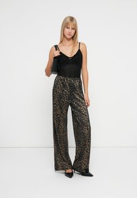 Body negru cu bretele subțiri, asociat cu pantaloni largi cu imprimeu leopard în culori aurii și negre, având o textură strălucitoare și talie înaltă. Ținând o geantă neagră.