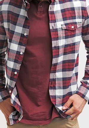 Personne portant une chemise à carreaux rouge, blanche et marine déboutonnée sur un polo rouge foncé, les mains reposant près des poches.