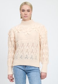 Maglione lavorato a maglia crema con colletto alto, dettagli floreali sulle spalle e un motivo a traforo con polsini e orlo a coste.