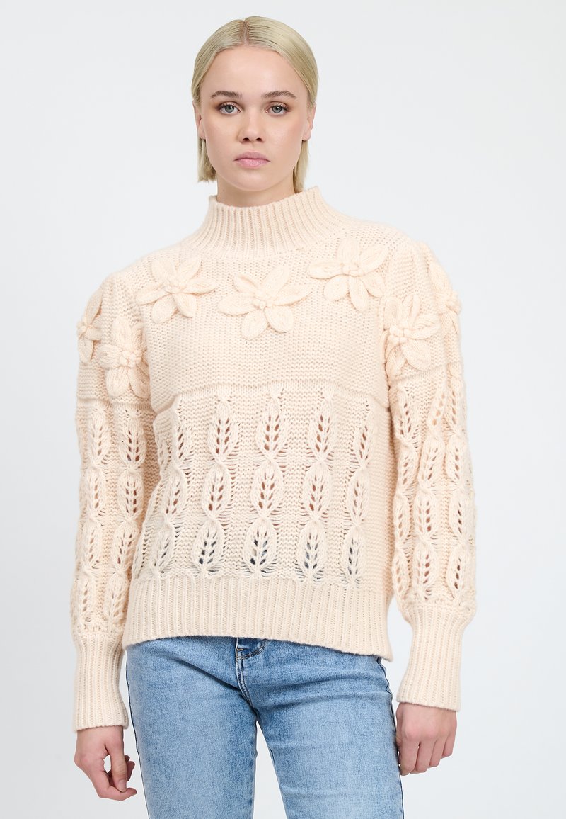Maglione lavorato a maglia crema con colletto alto, dettagli floreali sulle spalle e un motivo a traforo con polsini e orlo a coste.