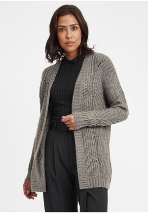 Cardigan en tricot gris avec un devant ouvert, texture côtelée, manches longues et fentes sur les côtés. Porté sur un haut noir et assorti à un pantalon noir.
