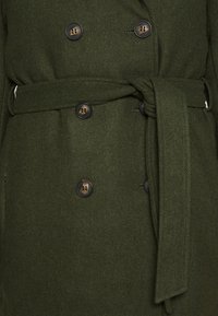 Abrigo verde de lana de doble botonadura con cinturón. Presenta botones marrones y una tela texturizada, creando una apariencia estructurada y con capas.