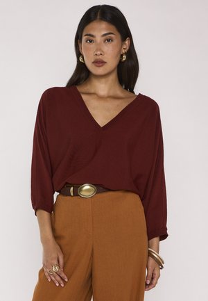 Giovane donna con lunghi capelli scuri che indossa orecchini dorati, una maglia bordeaux con scollo a V, pantaloni marroni a vita alta con cintura con fibbia dorata, anelli e braccialetti.