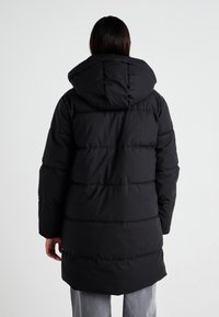 Chaqueta de abrigo negra con capucha, con secciones acolchadas y un corte de longitud media. La tela parece suave y mate, sin herrajes visibles.