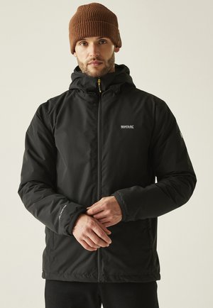 Homme portant une veste à capuche noire de la marque Regatta et un bonnet en tricot marron, ajustant ses manches, debout devant un fond clair uni.