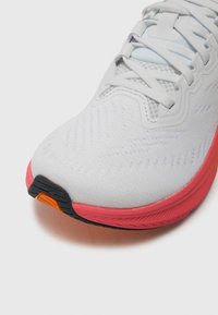 White athletic shoe with a textured upper and pink midsole. Features a black and orange outsole with a streamlined design and lace closure.