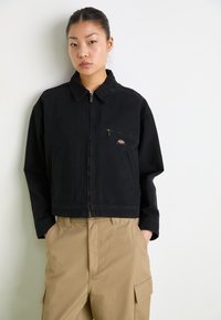 Zwarte cropped jack met een ritssluiting aan de voorkant, twee zijzakken en één borstzak. Voorzien van een kraag en een klein logo patch. Gladde textuur.