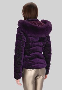 Fioletowa kurtka puchowa z kapturem obszytym sztucznym futrem, z pikowaną fakturą i błyszczącym wykończeniem. Noszona z metalicznymi złotymi spodniami.