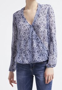 Blouse à motifs bleus et blancs en tissu léger, avec un col en V, des manches longues et des détails froncés à la taille.