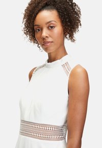 Vit ärmlös klänning med hög halsringning, utrustad med vävda detaljer vid axlarna och ett dekorativt utskuret band vid midjan.