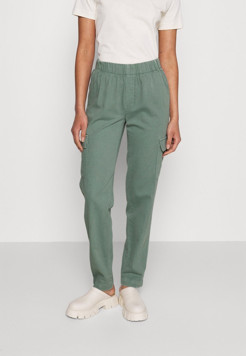 Pantalon cargo vert avec une taille élastique, des poches latérales et une coupe décontractée, associé à des sabots de couleur neutre et un t-shirt blanc uni.