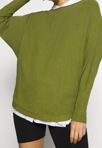 Pull en olive surdimensionné avec une texture côtelée, des manches larges et un ourlet arrondi, superposé sur une chemise blanche à col et un short noir.
