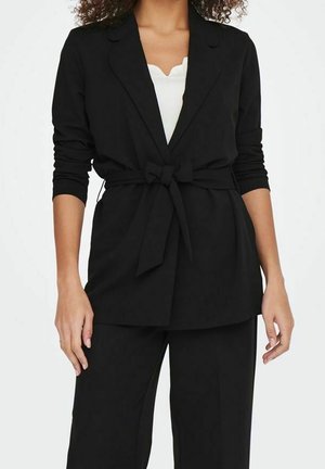 Woman wearing a black belted blazer over a white scalloped-edge top and matching black pants against a white background.