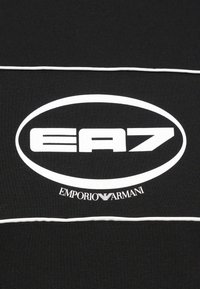 Melna T-kreklis, kas izgatavots no mīksta auduma, ar baltu EA7 logotipu ovālā un tekstu "EMPORIO ARMANI" apakšā. Ar baltu joslu kā akcentu.