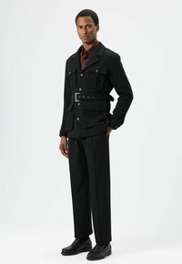 Zwarte ribfluwelen jas met meerdere zakken en een riem, gecombineerd met zwarte broek en schoenen. Model staat voor een effen achtergrond.