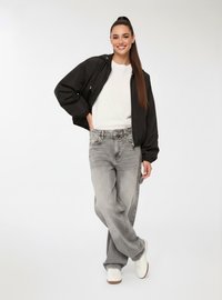 Giacca bomber nera, felpa bianca, jeans baggy grigi e sneakers bianche. Il modello sta in piedi con un piede incrociato, mani in tasca.