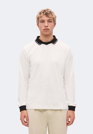 Un jeune homme se tient face à l’avant, vêtu d’un polo blanc à manches longues avec un col et des poignets noirs, ainsi que d’un pantalon beige.