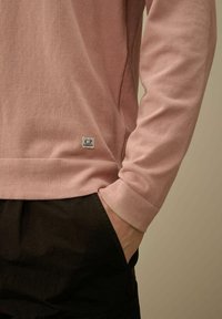 Camicia a maniche lunghe rosa chiaro con polsini a coste, che presenta un piccolo marchio C.P. Company sull'orlo inferiore. Indossata con pantaloni neri.