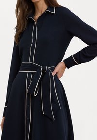 Vestido navy com detalhes em branco, fecho frontal com botões, decote com gola e faixa amarrada na cintura. Mangas longas e saia fluida com uma textura suave.