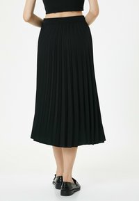 Zwarte geplooide rok met een elastische tailleband, gestructureerde stof en vloeiende A-lijnvorm. Draag met zwarte platte schoenen, weergegeven vanuit de achterkant.