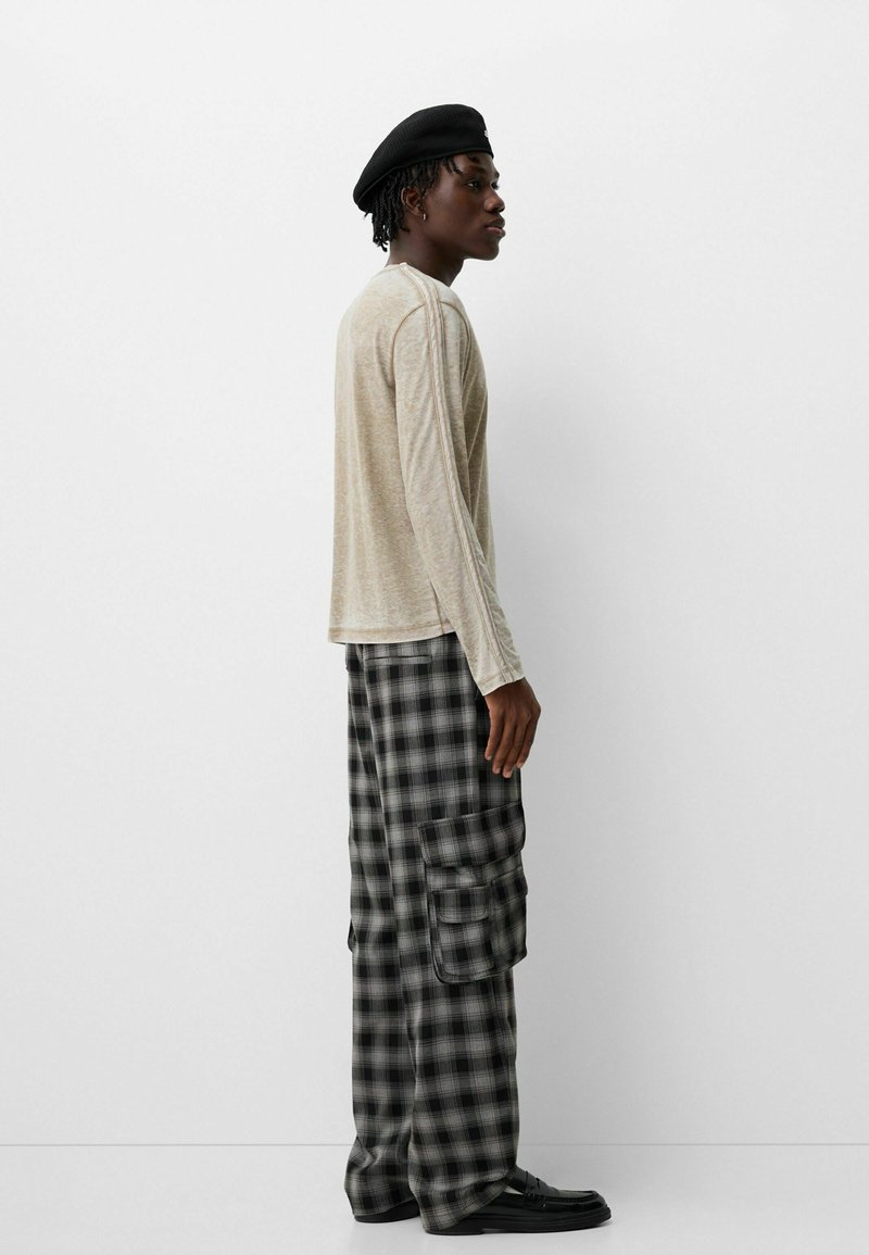 Camisa de manga larga color beige, pantalones cargo a cuadros en negro y gris, combinados con un boina negra y zapatos negros, vista de perfil contra un fondo blanco.