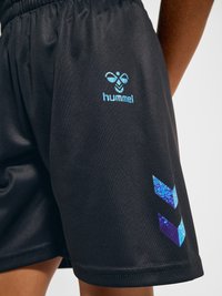 Schwarze Sportshorts mit blauem Hummel-Logo und Chevron-Mustern, getragen von einer Person mit sichtbarer brauner Haut an Bein und Arm.