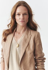 Blaser en lin beige sur un haut en maille beige, avec un collier en pendentif doré. Cheveux ondulés doux, fond neutre.
