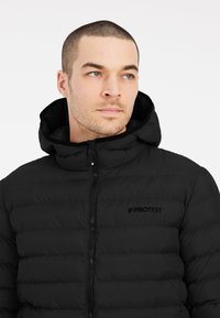 Zwarte pufferjack met een ritssluiting aan de voorkant, hoge kraag en capuchon. Beschikt over een gequilt ontwerp en een discreet logo op de borst.