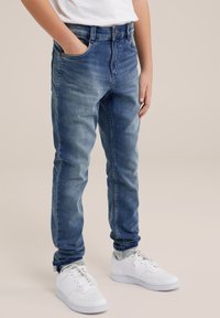 Jeans in denim blu con un design aderente, tasca frontale singola e leggera scoloritura. Abbinati a sneakers bianche, con chiusura a lacci e texture liscia.