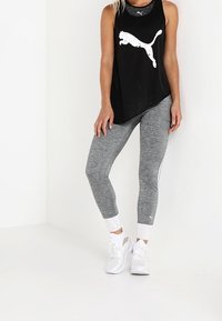 Kobieta w czarnej koszulce na ramiączkach Puma, szarych legginsach z białymi paskami, białych skarpetkach i białych butach sportowych, stojąca na tle białej ściany.