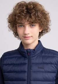 Gilet rembourré bleu marine avec motif matelassé, col haut et fermeture éclair à l'avant. Il présente une petite écusson logo sur le côté gauche. Texture lisse.
