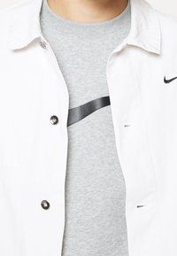 Person som bär en ljusgrå Nike T-shirt under en öppen vit skjorta med knappar och en liten svart Nike swoosh-logga.