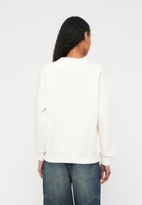 Sahnefarbener Sweatshirt mit lockerer Passform, gerippten Bündchen und Saum. Getragen mit blauen weitem Jeanshosen, von hinten vor hellem Hintergrund gezeigt.