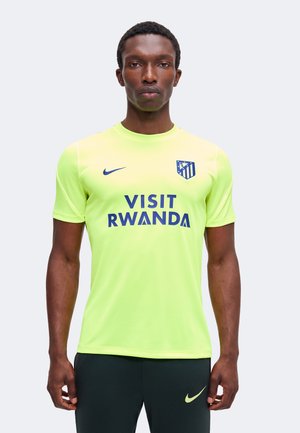 Hombre vistiendo una camiseta de fútbol del Atlético de Madrid en amarillo neón con los logotipos de "Visit Rwanda" y Nike, posando frente a un fondo claro y liso.