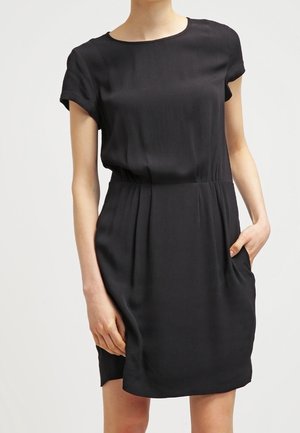 Woman wearing a simple black short-sleeve dress with a round neckline and one hand in a side pocket.