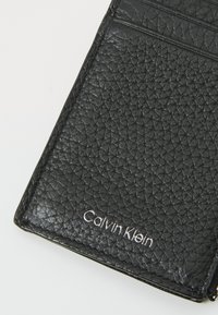 Czarny skórzany portfel z teksturowanym wykończeniem, z wieloma kieszonkami na karty i srebrnym logo "Calvin Klein" z przodu.