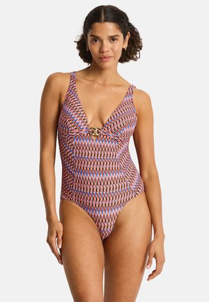 Frau trägt einen bunten Badeanzug mit geometrischem Muster und einem goldenen Verschluss in der Mitte vorne, steht vor einem neutralen Hintergrund.