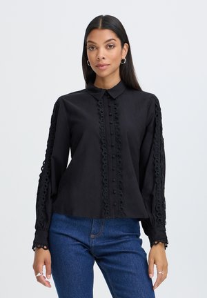 Vrouw met lang donker haar draagt een zwarte blouse met knopen en kanten afwerking op mouwen en voorkant, gecombineerd met een blauwe spijkerbroek, staand tegen een lichte achtergrond.