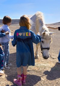 Veste en jean avec des lettres colorées "POWER" et des franges, portée par un enfant près d'un cheval blanc ; bottes à franges roses ; cadre extérieur.