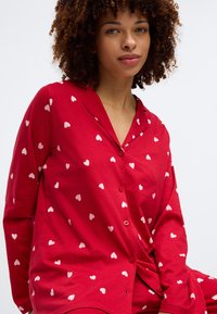 Κόκκινη πιτζάμα με κουμπιά, με λευκά σχέδια καρδιών. Κατασκευασμένη από μαλακό ύφασμα, έχει χαλαρή εφαρμογή και γιακά.