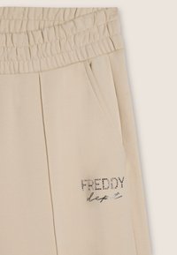 Pantalones deportivos beige con cintura elástica, bolsillos laterales y logo "FREDDY" en strass. Tejido suave con una textura lisa.