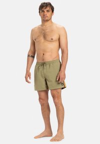 Zelene kopalne kratke hlače z zategovalnim trakom v pasu, stranskimi črtami v rdeči in rumeni barvi ter logotipom na robu. Gladek, lahek material.