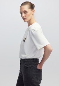Witte T-shirt met korte mouwen en een ronde halslijn, voorzien van een decoratieve kraaldecoratie op de borst, gecombineerd met zwarte high-waisted jeans.