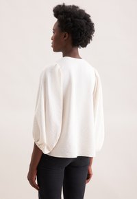 Hvid tekstureret bluse med pufærmer, rund halsudskæring og afslappet pasform. Kombineret med mørke jeans, set fra ryggen.