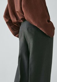 Persona che indossa una camicia marrone a maniche lunghe ampia infilata in pantaloni grigi scuri a gamba larga a vita alta su sfondo chiaro.