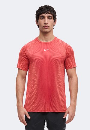 Giovane uomo che indossa una maglietta sportiva rossa Nike a maniche corte con logo swoosh bianco, in piedi di fronte a uno sfondo uniforme.