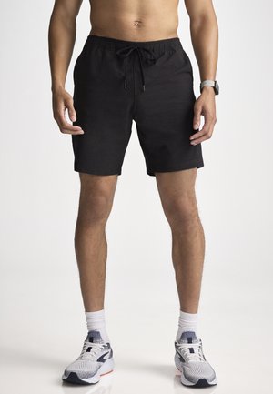 Mann trägt schwarze Shorts mit Kordelzug, weiße Socken und weiße Turnschuhe vor einfarbigem weißem Hintergrund, dargestellt von der Taille bis zu den Füßen.
