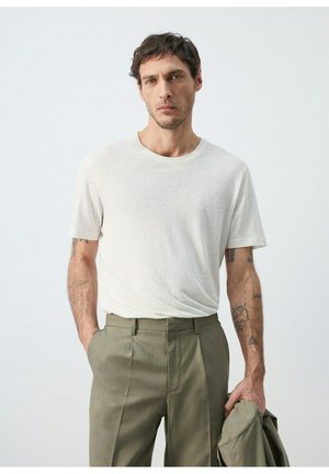 Uomo con capelli corti e ricci, indossa una maglietta bianca e pantaloni color oliva, tiene una giacca oliva nella mano sinistra, in piedi davanti a uno sfondo semplice.