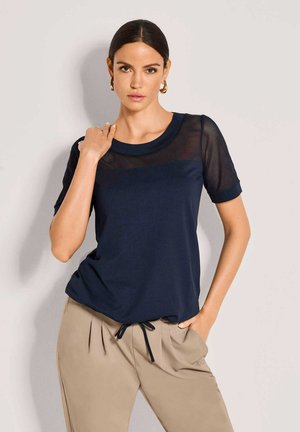 Vrouw draagt een marineblauw gaastopje en beige broek, staat met één hand in de zak en de andere de kraag aanrakend, tegen een effen achtergrond.