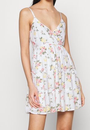 Vrouw die een mouwloze witte jurk met bloemenprint draagt, met roze en gele bloemen en een gelaagde rok, staand tegen een effen achtergrond.